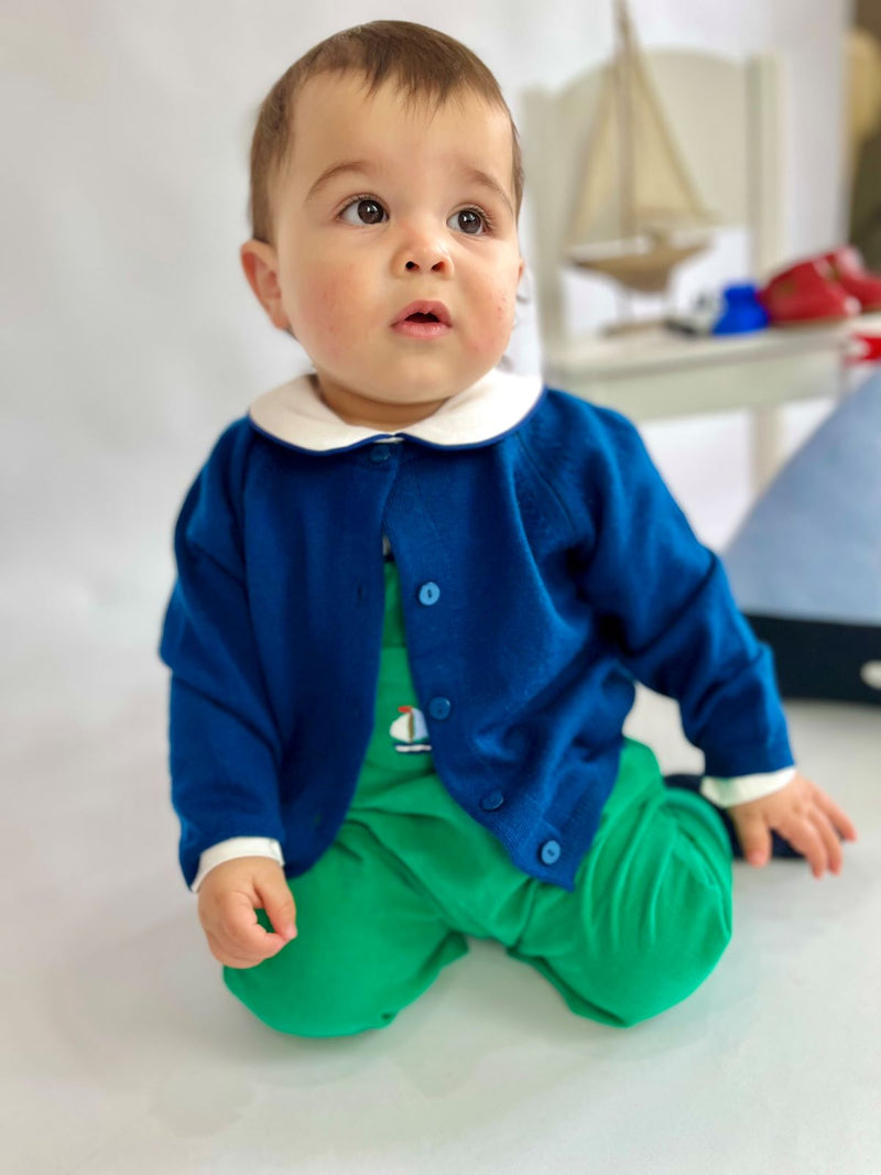 Child wearing green corduroy overalls matched with blue cardigan and white bodysuit featuring blue piping on the collar.