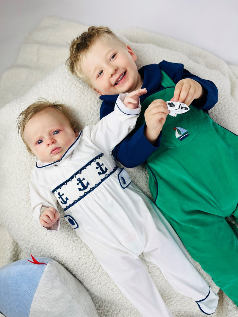 Child wearing green corduroy overalls matched with a blue classic jersey top laying next to a baby wearing a nautical anchor babygrow.